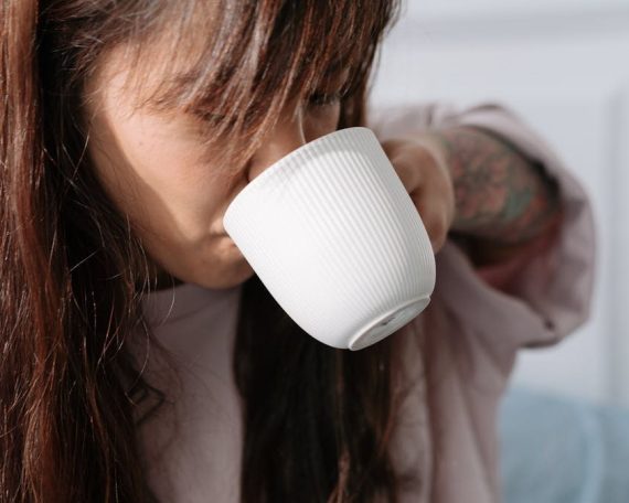 A woman with long hair sipping coffee from a white cup inside a cozy room.